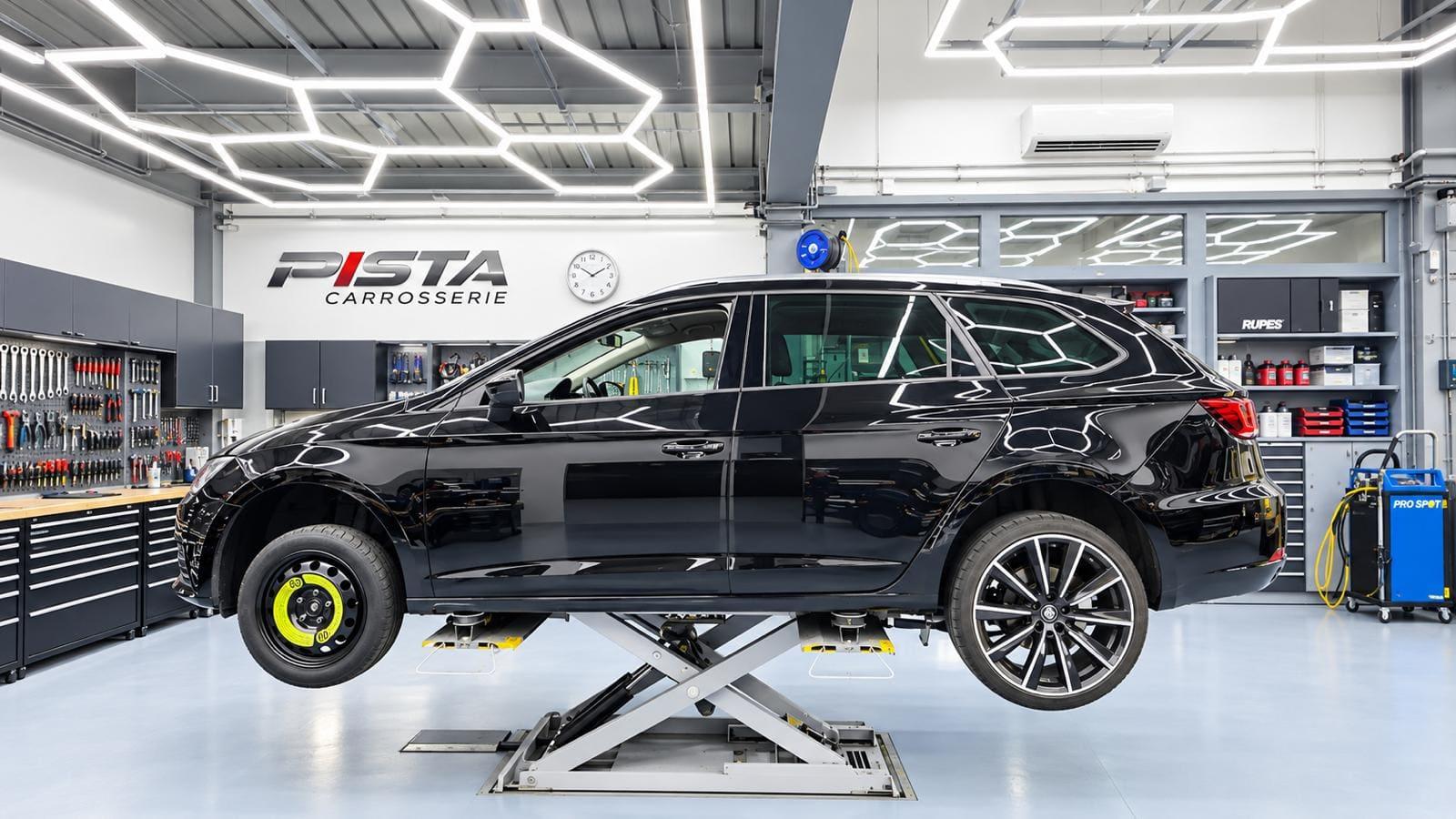 Atelier de Carrosserie Pista : un break noir sur pont élévateur, établis d'outillage, éclairage LED hexagonal et logo de l'entreprise au mur